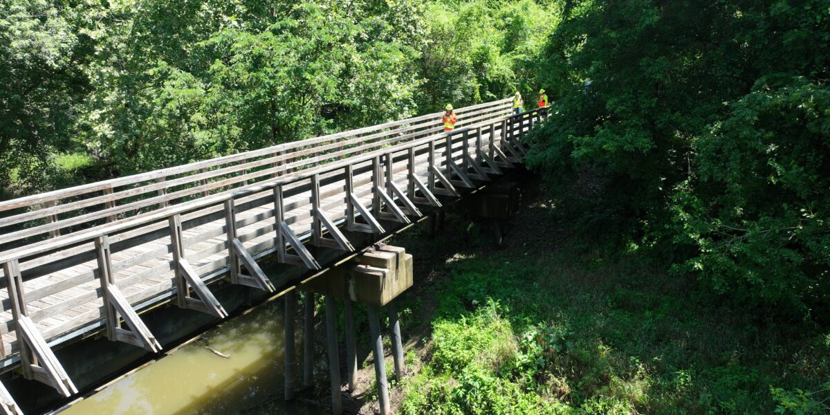 Close to ground-level view of a trail bridge over water, with Horner & Shifrin engineers in PPE conducting a structural inspection, surrounded by trees and green vegetation.