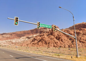 Traffic signal at the US-191 and Arches National Park entrance intersection in Grand County, Utah, with the Arches National Park road sign visible, red rock cliffs in the background, and a motorcyclist and vehicle traveling along the highway.