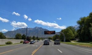 Illuminated variable message sign over the roadway displaying “Little Cottonwood Canyon” and a 24-minute travel time, with a mountain backdrop, green landscape, and light blue sky in Cottonwood Heights, Utah.