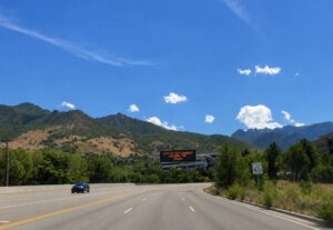 Illuminated variable message sign over the roadway displaying “Little Cottonwood Canyon” and a 25-minute travel time in yellow text on a black sign, with a 50 mph speed limit sign visible to the right, green hills in the background, and trees lining both sides of the road.