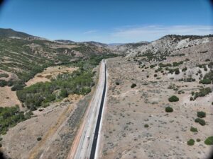 Aerial view of SR-132 in Sanpete County, Utah, showing a four-lane highway with vehicles traveling in both directions, fresh asphalt on the right shoulder, construction cones visible, a curve ahead at the horizon, desert terrain with scattered trees and bushes, power lines to the right, and mountains in the background.