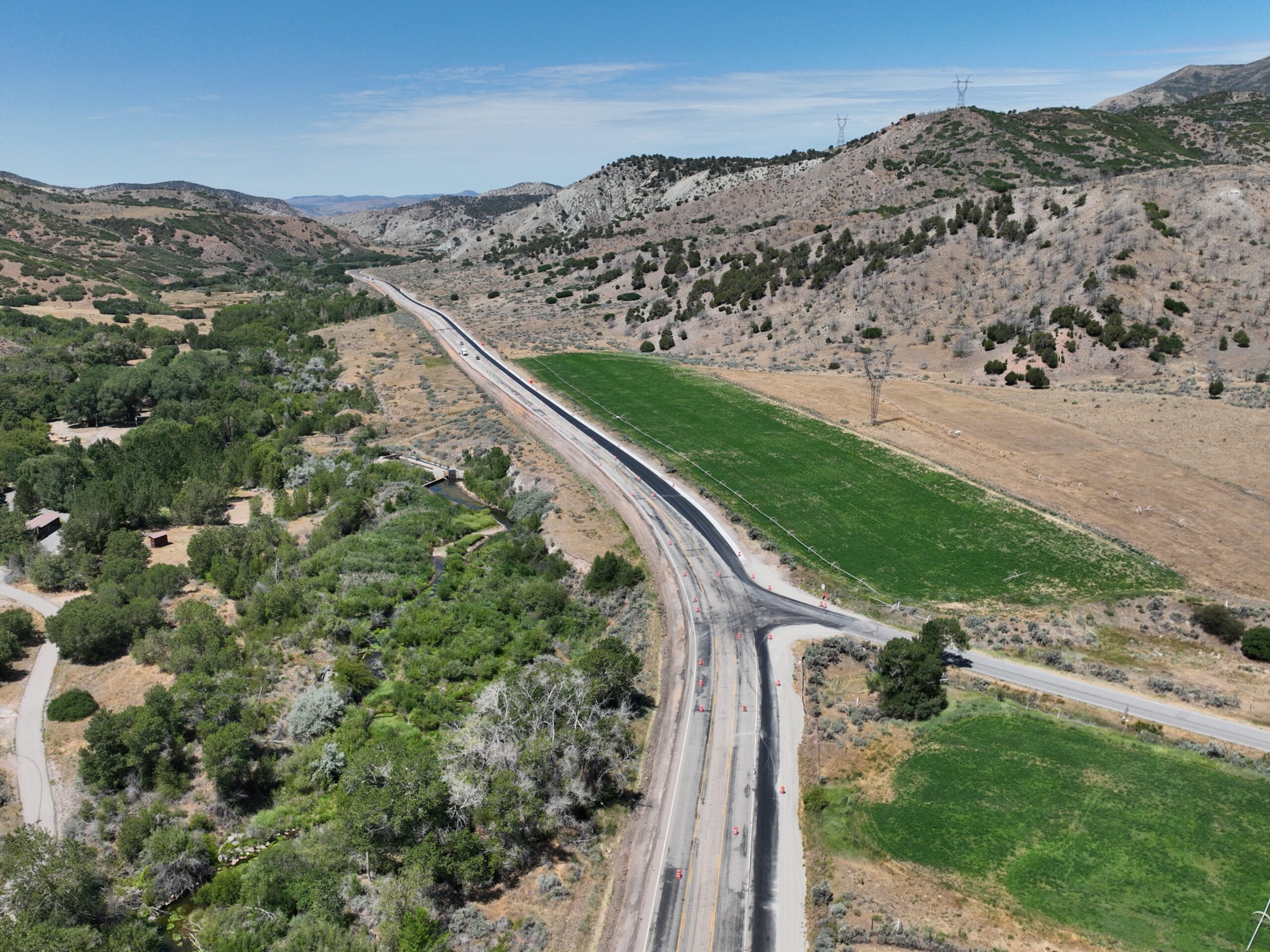 Aerial view of SR-132 near the Nebo Loop Road intersection in Sanpete County, Utah, showing the road forking with a grassy median and a single large tree at the split, rolling green hills, scattered houses and a small outbuilding visible among trees to the left, and sectioned green fields to the right.