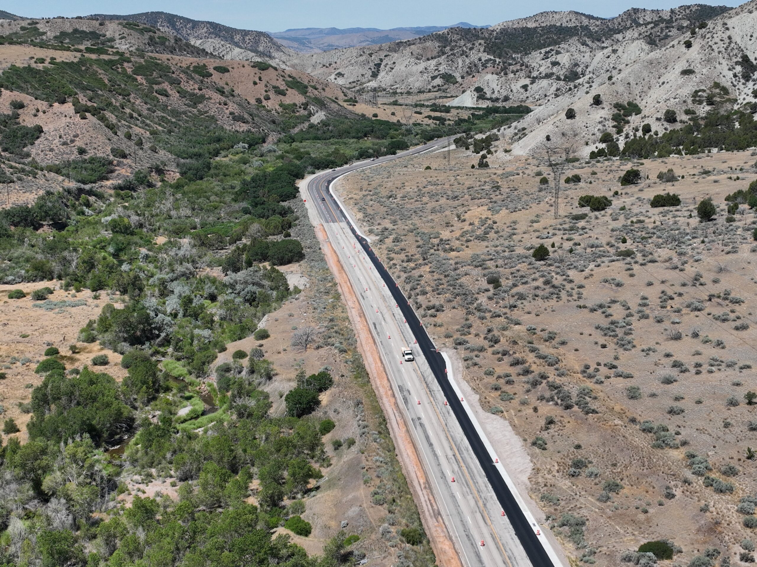 Wide aerial drone shot of SR-132 in Sanpete County, Utah, showing the highway bending toward the horizon with rolling mountains and hills extending into the distance, desert terrain transitioning to green hills, and the full corridor visible from above.