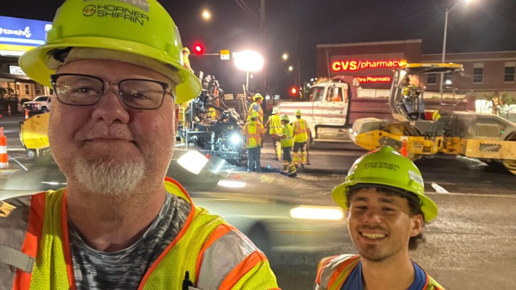 Sloan Smith and Adam Bier smiling in hard hats on a night construction job with a crew working in the background.
