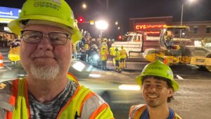 Sloan Smith and Adam Bier smiling in hard hats on a night construction job with a crew working in the background.