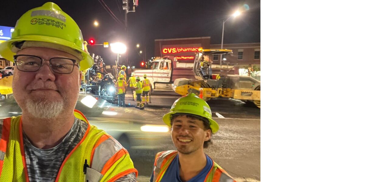 Sloan Smith and Adam Bier on a night construction job, smiling in hard hats with a construction crew working in the background.