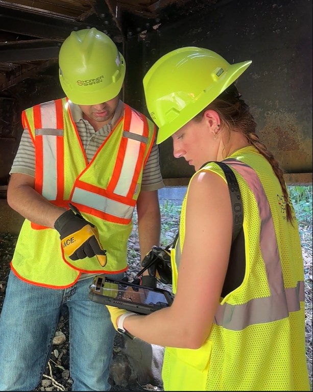 Utkarsh Kode and Norah Gum under the MCT Trail bridge during a structural inspection, with Norah entering data on a tablet.