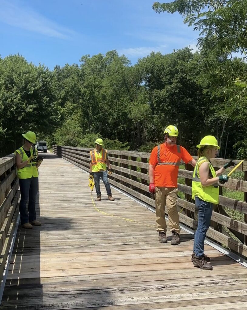 Sara Rogers, Norah Gum, Utkarsh Kode, and Tom Lohman on the MCT Trail bridge during a structural inspection, all in full PPE including high-visibility vests and hard hats.