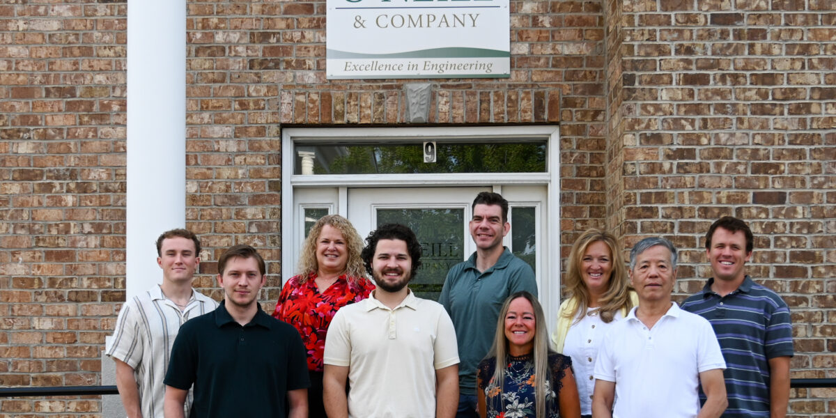 The O’Neill & Company team of nine poses together outside their Salt Lake City office, smiling in front of the building entrance with O’Neill & Company signage visible above and on the glass door behind them.