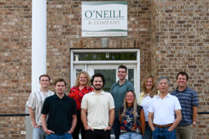 The O’Neill & Company team of nine poses together outside their Salt Lake City office, smiling in front of the building entrance with O’Neill & Company signage visible above and on the glass door behind them.