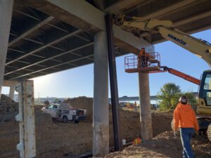 Construction crew working around vertical steel H-piles and a yellow excavator beneath a bridge pier cap