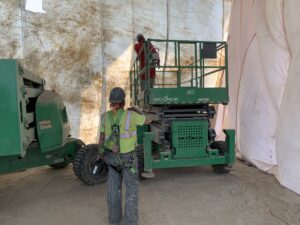 Two workers in a sealed bridge painting containment structure, one on a Skyjack scissor lift and one on the ground wearing a safety harness