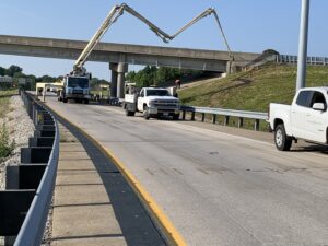 Horner and Shifrin branded pickup truck parked on a highway ramp shoulder with a concrete pump truck extending its boom over the bridge in the background