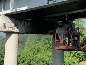 Crew Member in white protective suit using a lift basket to blast or clean a heavily damaged concrete pier cap with exposed rebar