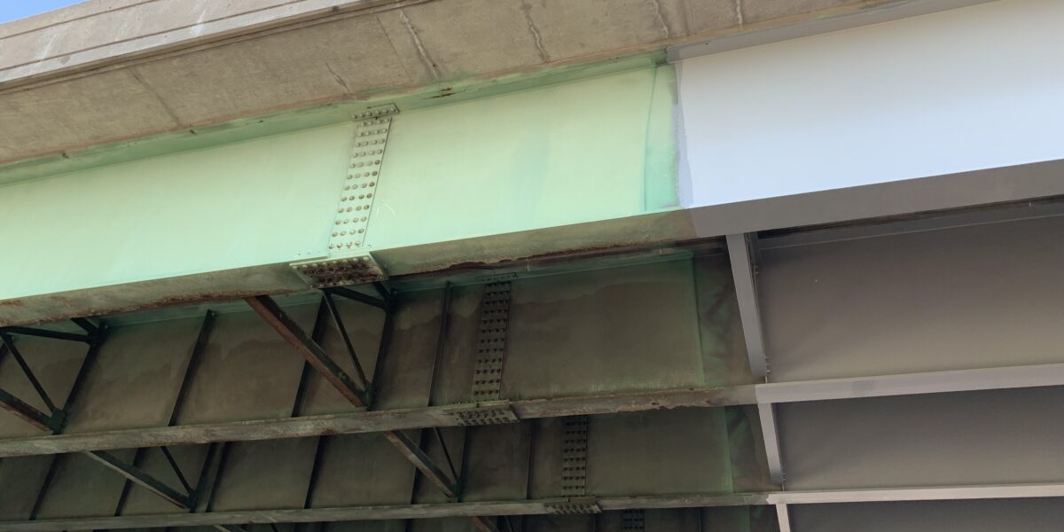 Underside of a highway bridge showing weathered green rusted steel girders on the left next to a freshly painted light-gray girder on the right