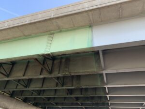 Underside of a highway bridge showing weathered green rusted steel girders on the left next to a freshly painted light-gray girder on the right