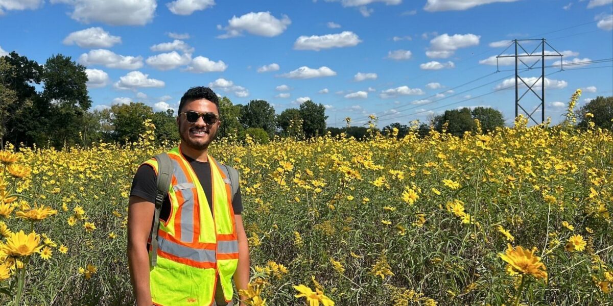 Xavier Diggs smiling outdoors in a sunflower field, wearing a reflective safety vest and backpack.