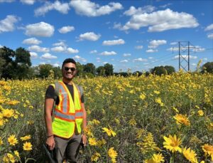 Xavier Diggs smiling outdoors in a sunflower field, wearing a reflective safety vest and backpack.