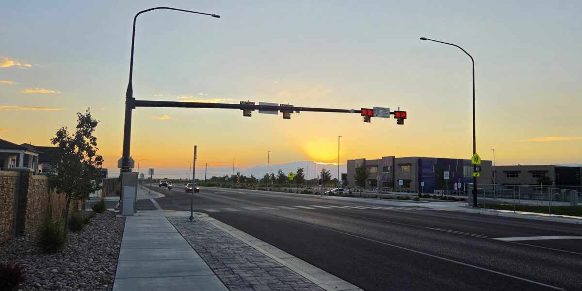 Overhead traffic signal mast arm with red signal heads and a school crosswalk warning sign above SR-193 in West Point, Utah, at sunset.